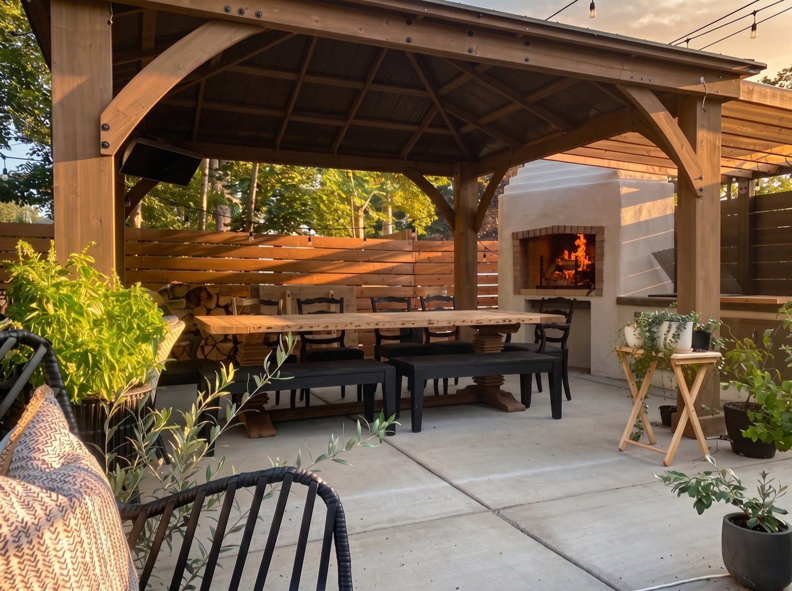 Timber pergola pavilion with a long reclaimed-wood dining table and a lit wood-fired fireplace at golden hour